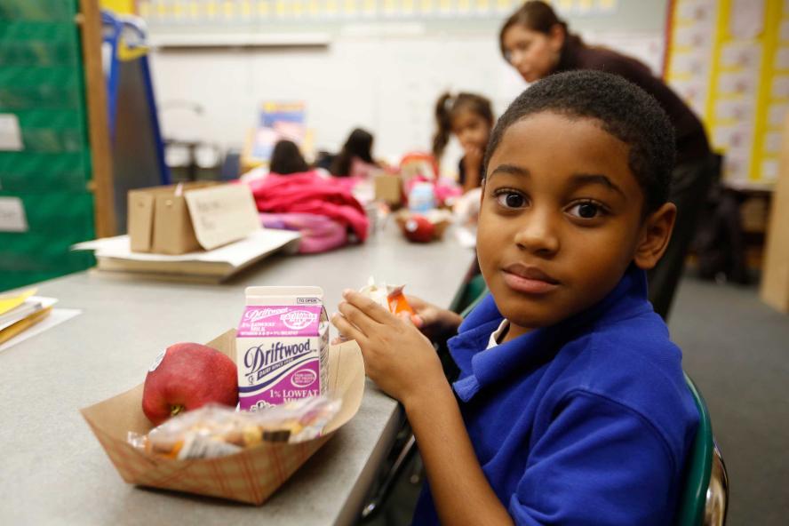 A boy in a blue shirt sitting and looking at the camera while eating breakfast in a school setting