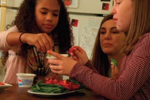 Professor Folta observing as one kid holds a dip container and another dips a vegetable, with more vegetables on a plate on the table