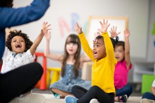 Kids sitting on the floor with their teacher, raising both hands and smiling, as part of the Active Start Project