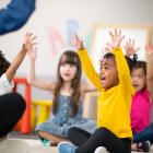 Kids sitting on the floor with their teacher, raising both hands and smiling, as part of the Active Start Project